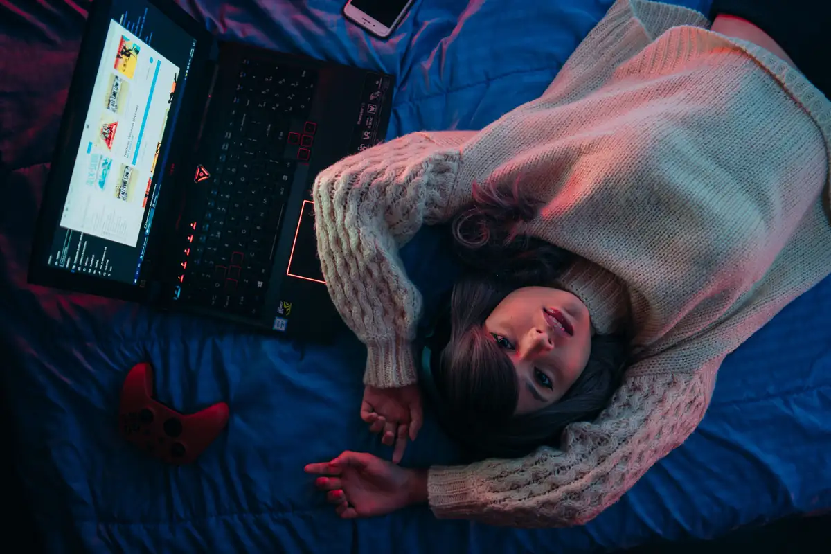 A person lying on a bed next to an open gaming laptop and a game controller